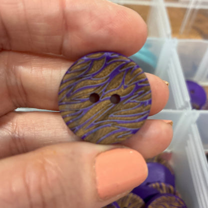 Hand holding a purple and gold button with a container of buttons in the background