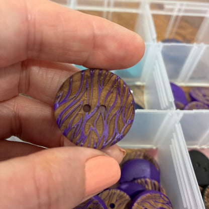 Hand holding a decorative button with purple and gold pattern, with a container of similar buttons in the background.