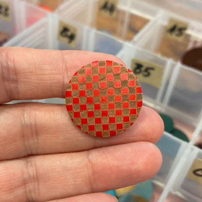 Hand holding a red and gold checkered button with a container of similar buttons in the background