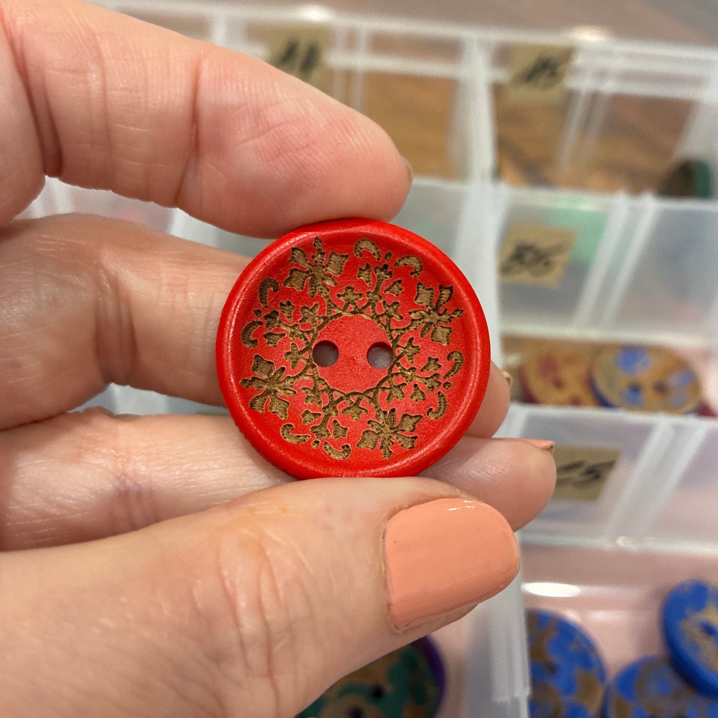 Hand holding a red decorative button with intricate design, against a blurred background of a storage box with more buttons.