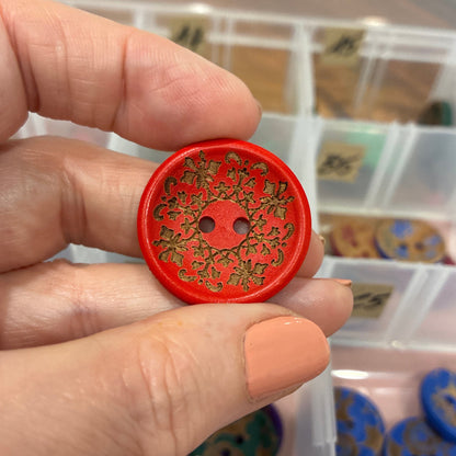 Hand holding a red decorative button with intricate design, against a blurred background of a storage box with more buttons.