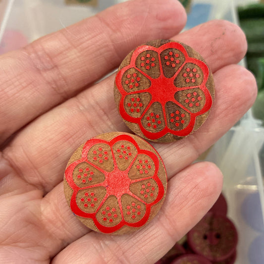Hand holding two red floral buttons with a container of assorted buttons in the background.