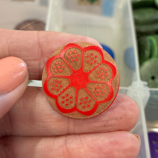 Hand holding a red floral button with a background of various buttons in a container.