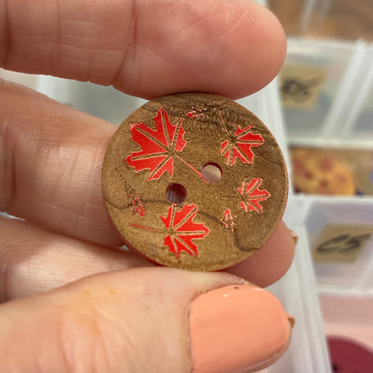 Hand holding a decorative button with red floral patterns against a blurred background of a container with various buttons.