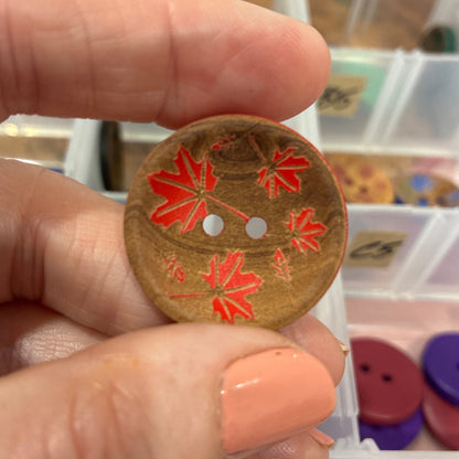 Hand holding a decorative button with red leaf patterns against a background of button containers.