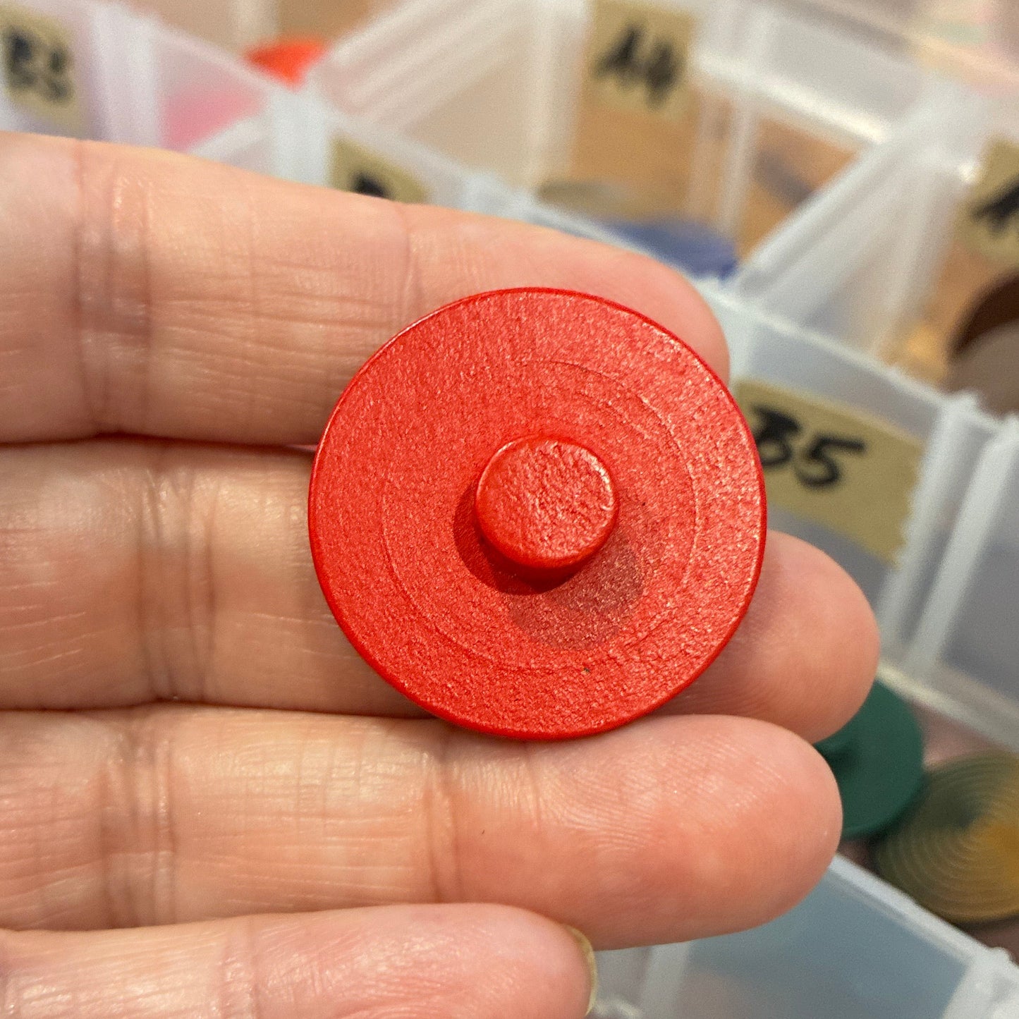Red circular button held in a hand with a container of colorful round objects in the background