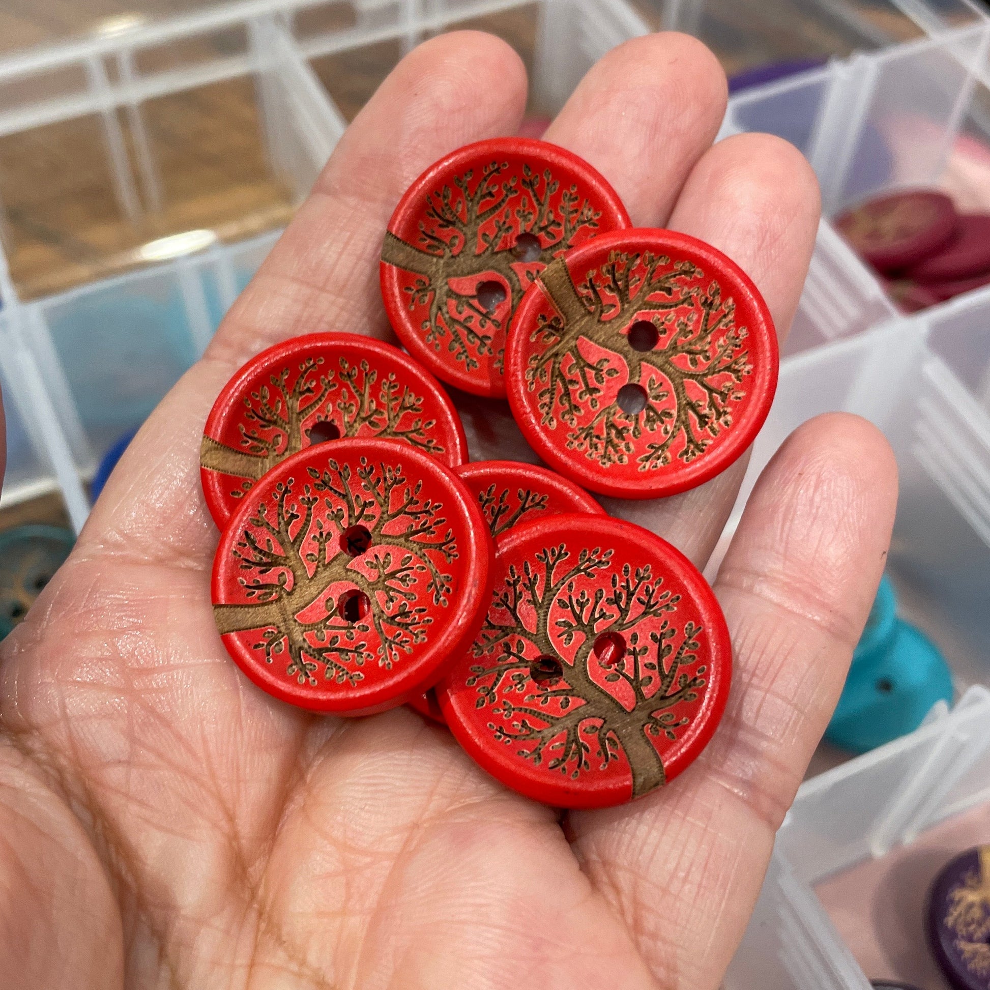 Hand holding red buttons with tree design against a blurred background of button containers.