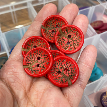 Hand holding red buttons with tree design against a blurred background of button containers.