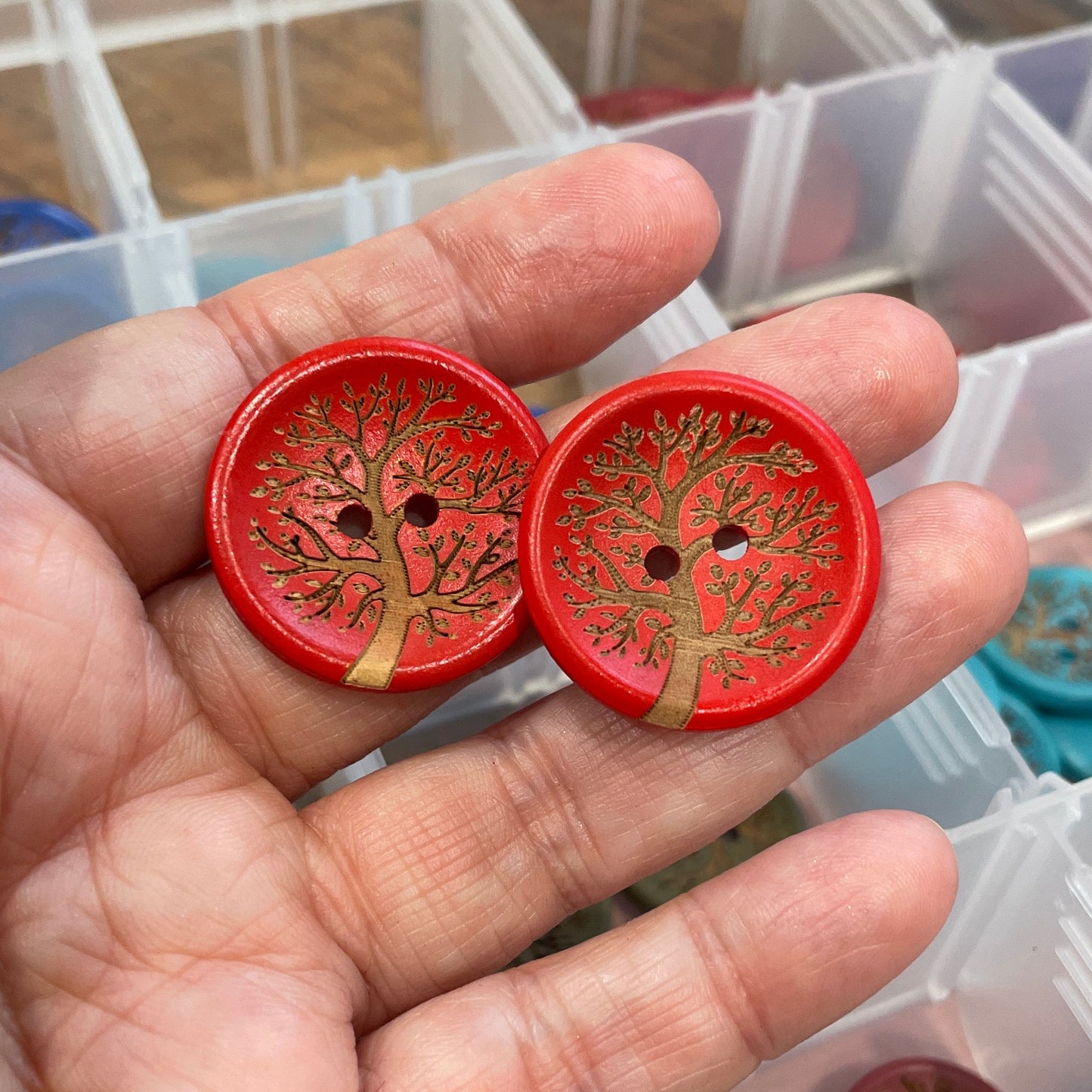 Hand holding two red buttons with intricate designs, with a container of similar buttons in the background.