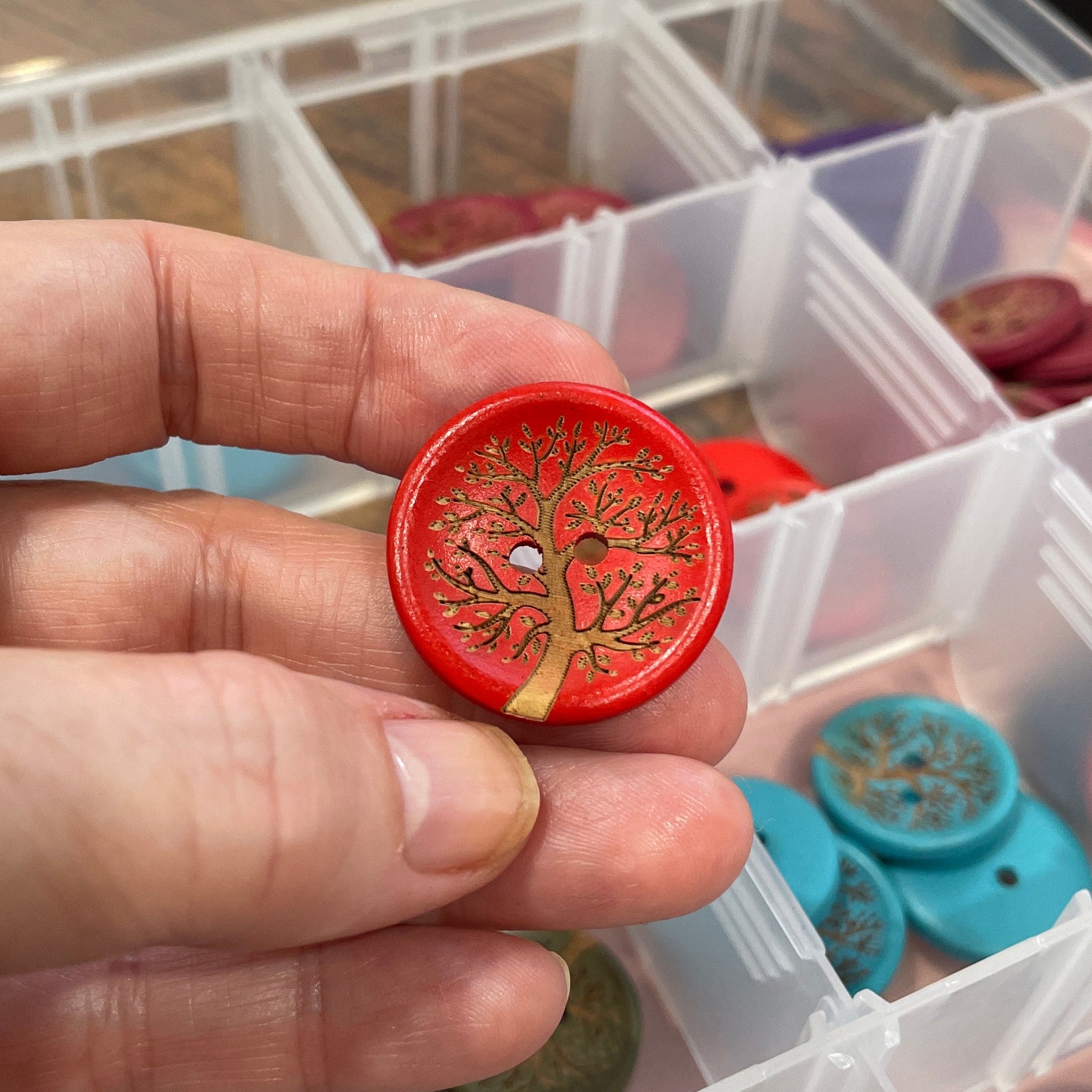 Hand holding a red decorative button with a tree design, against a background of a clear container with various buttons.
