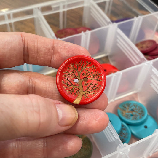Hand holding a red decorative button with a tree design, against a background of a clear container with various buttons.