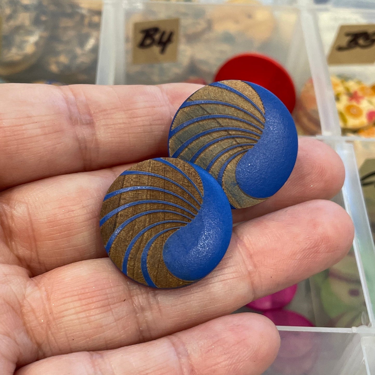 Hand holding two blue buttons with gold patterns in front of a container of various buttons.