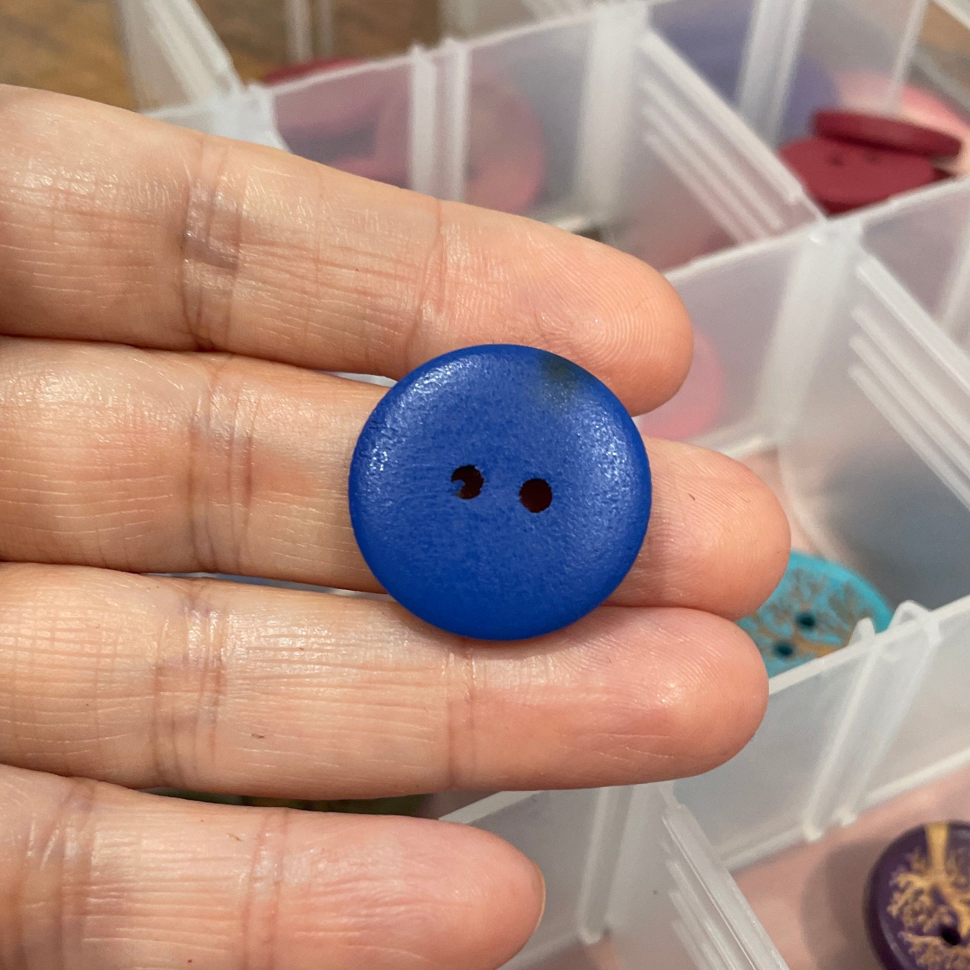 Hand holding a blue button with a background of assorted buttons in a clear container.