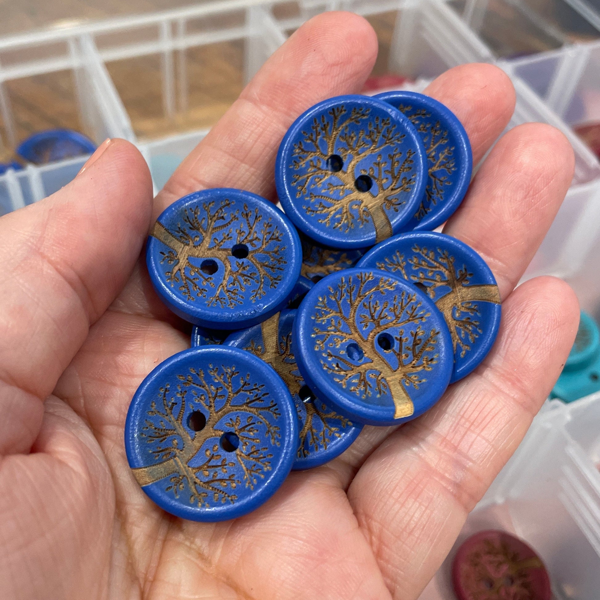 Hand holding blue buttons with gold tree designs over a container of similar buttons.