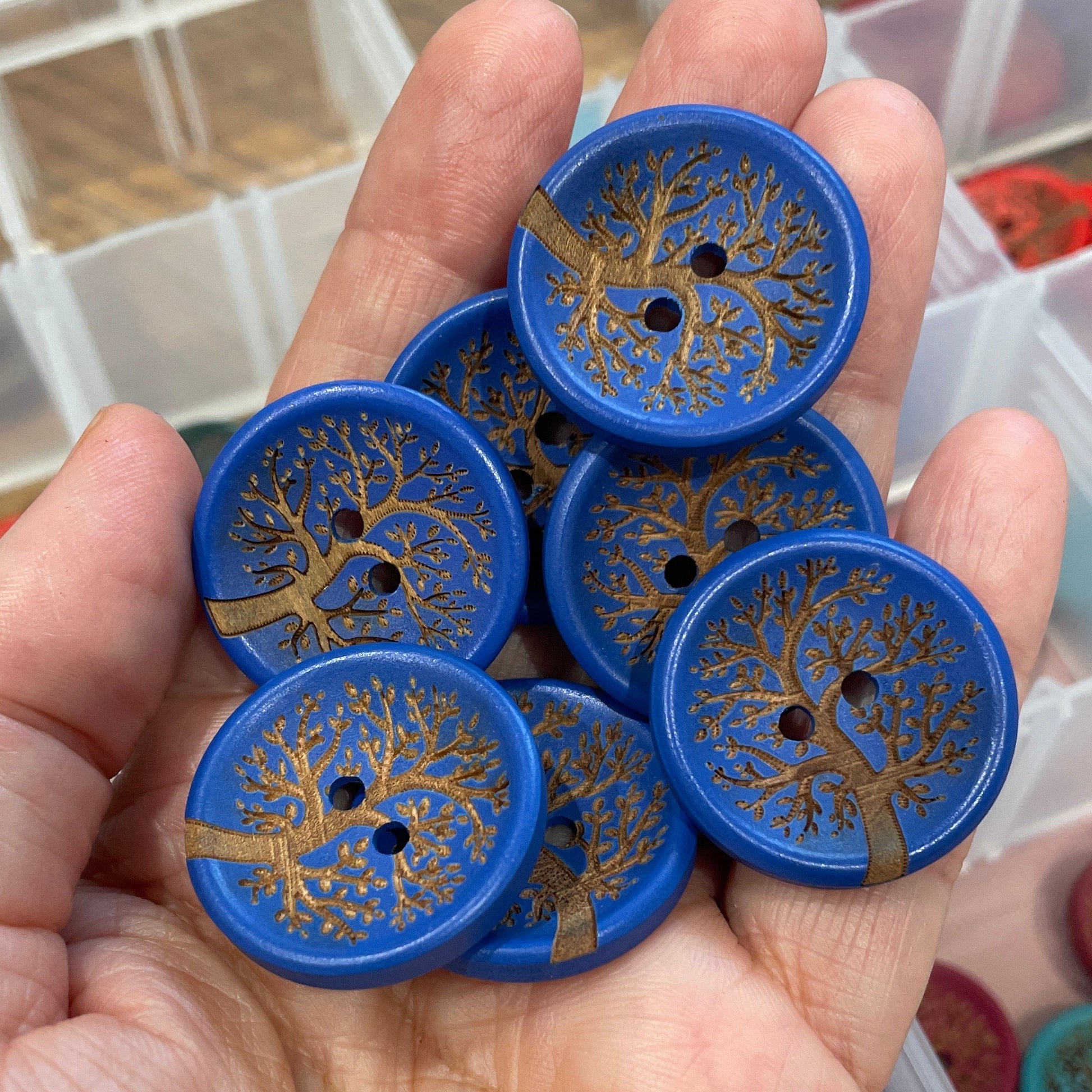 Hand holding blue buttons with tree design in front of a clear container.