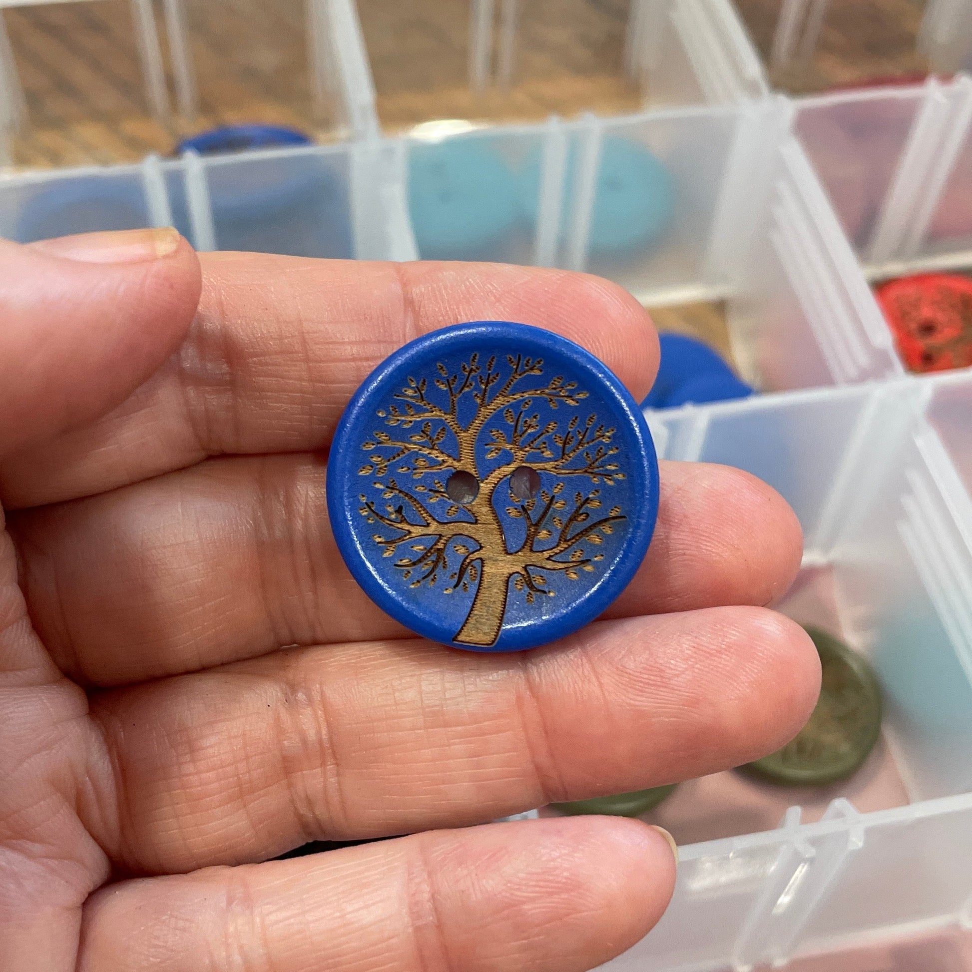 Hand holding a blue button with a tree design in front of a container of buttons.