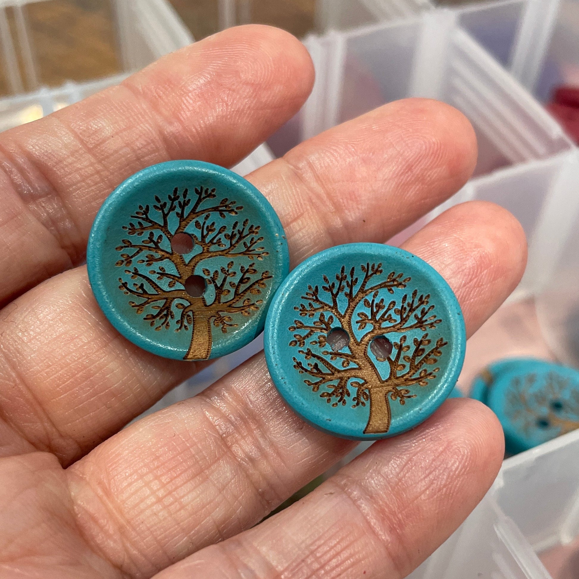 Hand holding two blue buttons with tree design in front of a clear container.