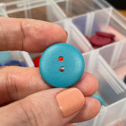 Hand holding a blue button with a clear plastic container of buttons in the background