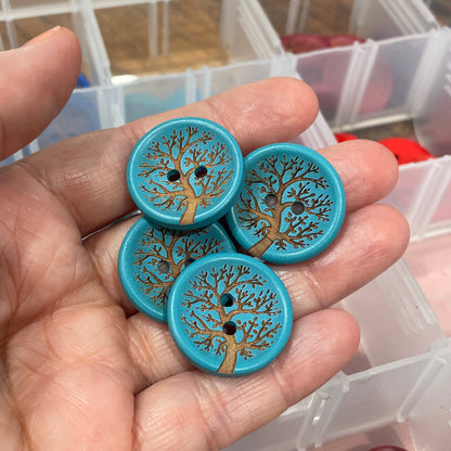 Hand holding decorative buttons with tree design, with a container of similar buttons in the background.