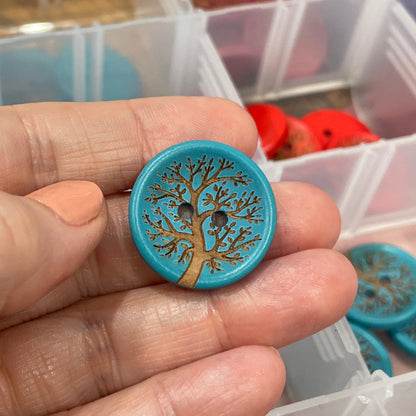 Hand holding a decorative button with a tree design, with a container of buttons in the background.