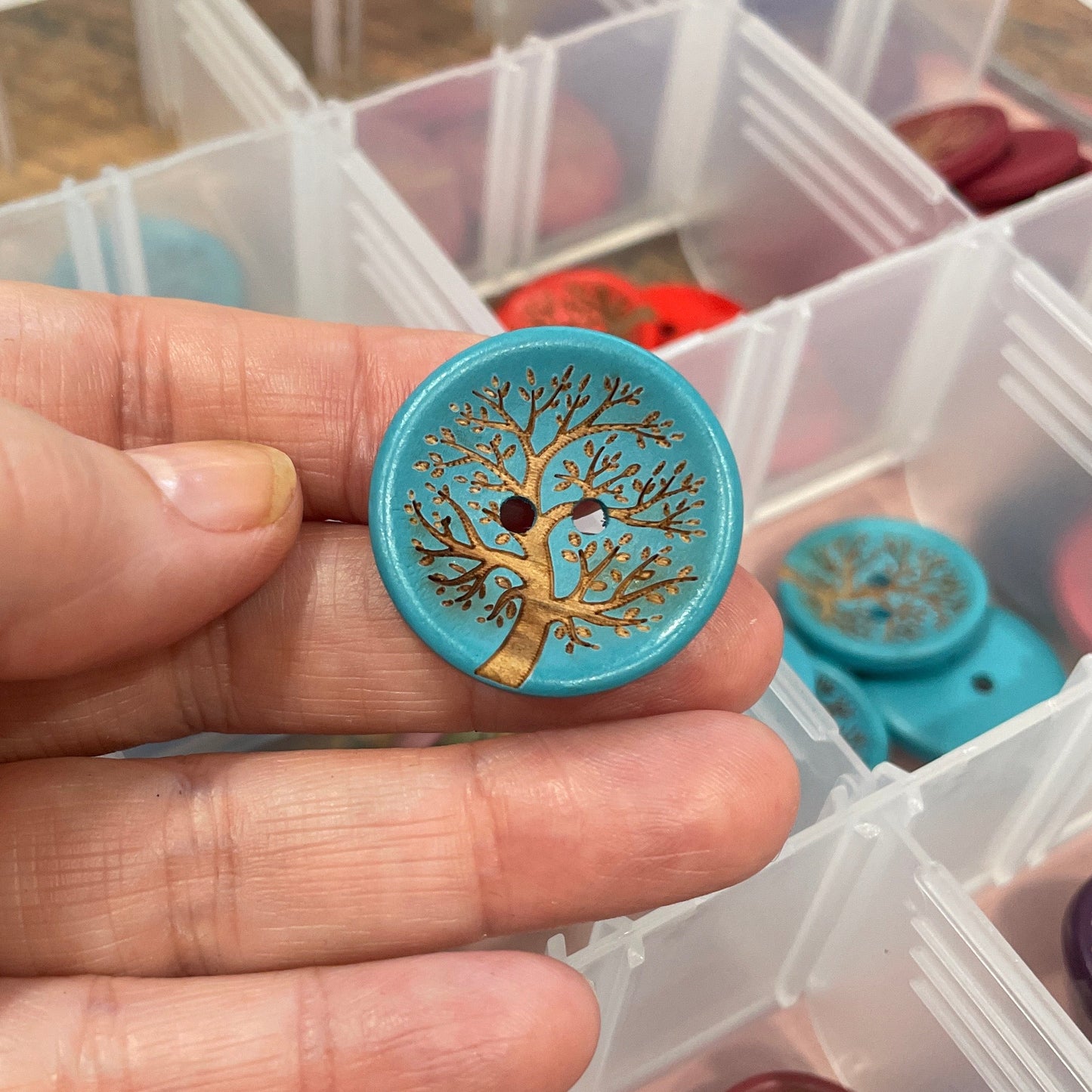 Hand holding a decorative button with intricate design, surrounded by more buttons in a clear container.