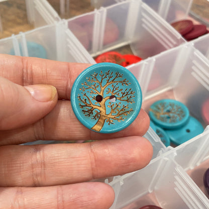 Hand holding a decorative button with intricate design, surrounded by more buttons in a clear container.