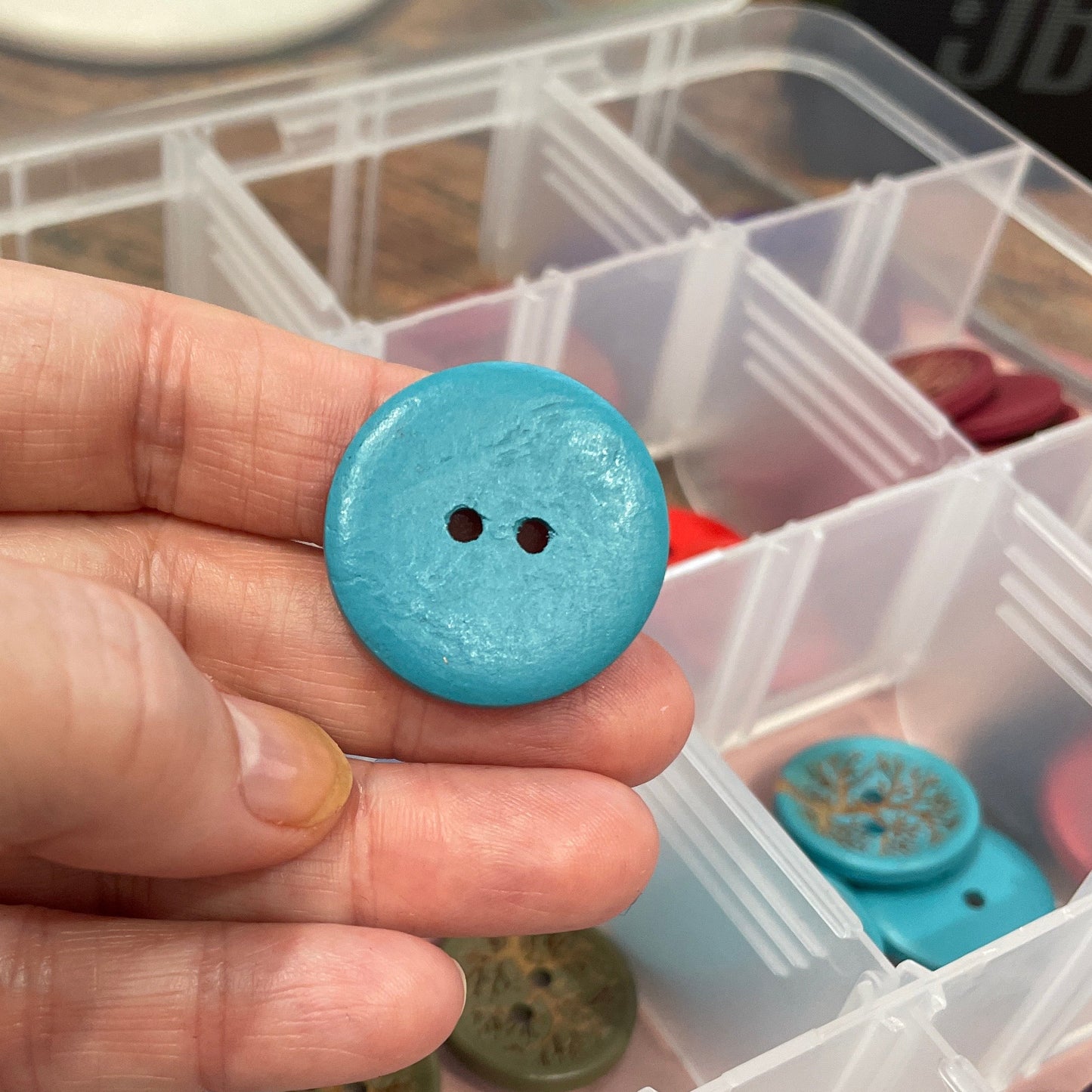 Hand holding a blue button with a container of various buttons in the background