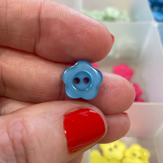 Hand holding a blue flower-shaped button with a container of colorful buttons in the background.