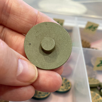 Hand holding a round green button with a clear container of similar buttons in the background.