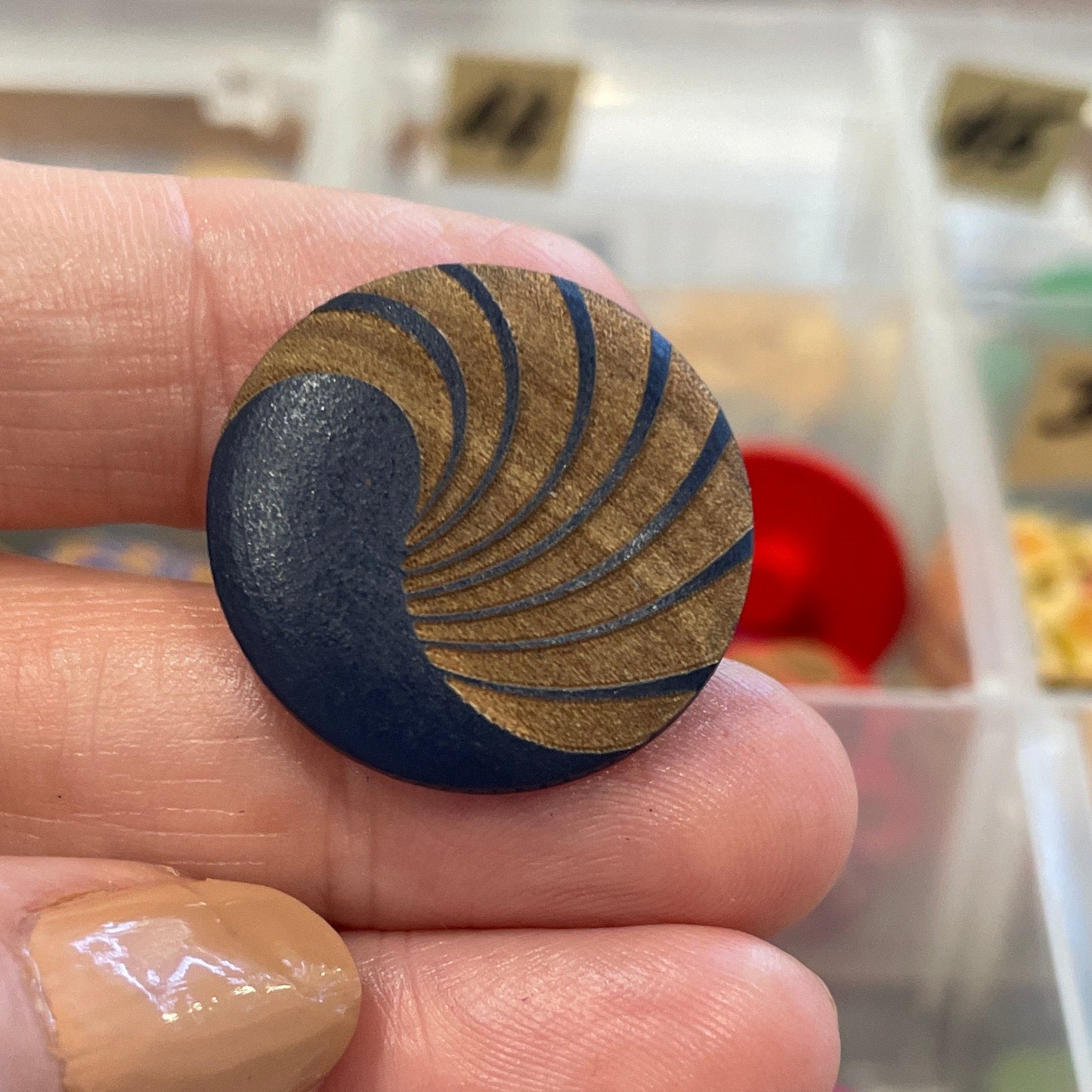 Hand holding a black and gold patterned button with a colorful button collection in the background.