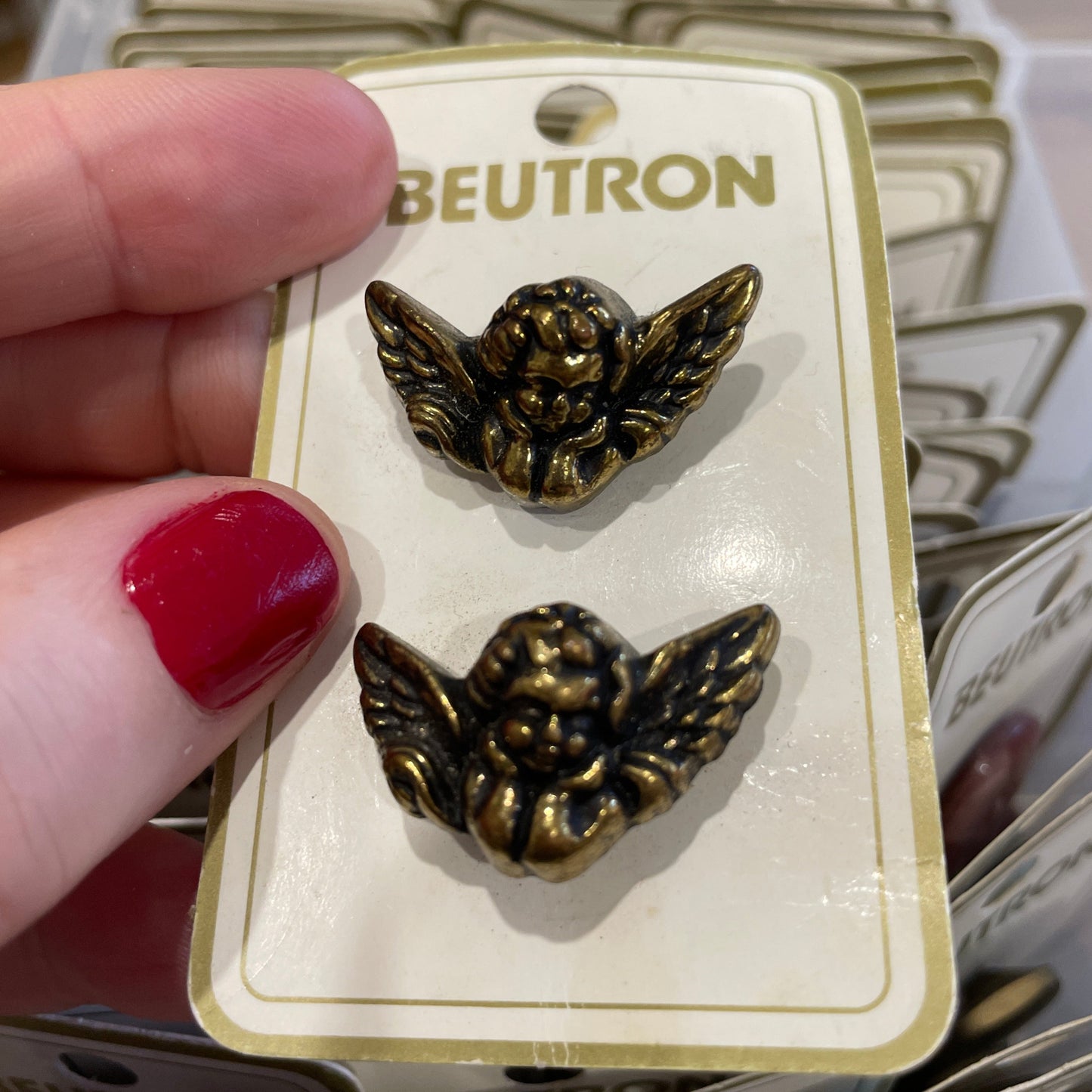 Gold winged buttons on a Beutron card held by a hand with red nail polish.