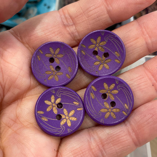 Purple buttons with floral designs held in a hand, with a container of buttons in the background.