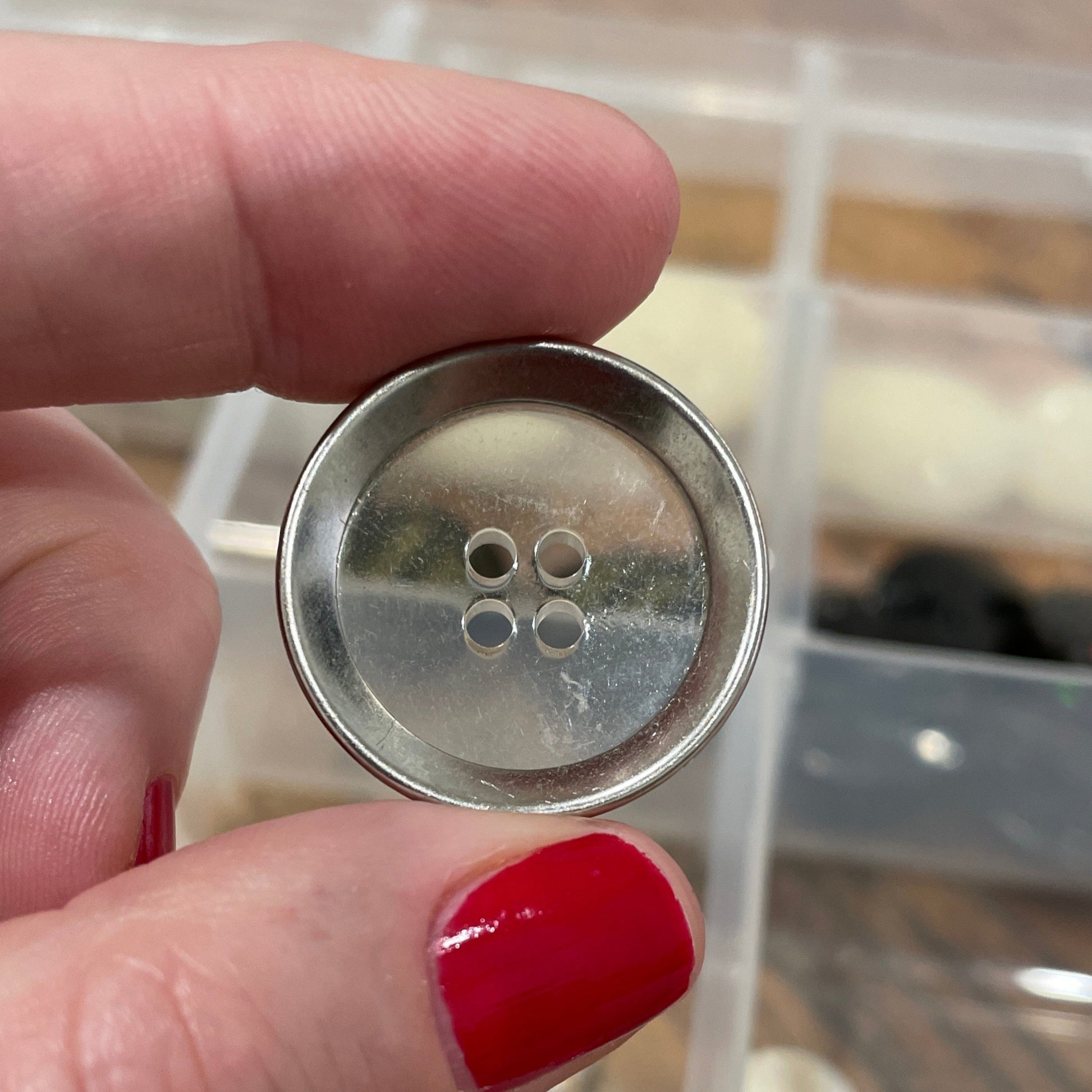 Hand holding a metal button with a clear container of buttons in the background