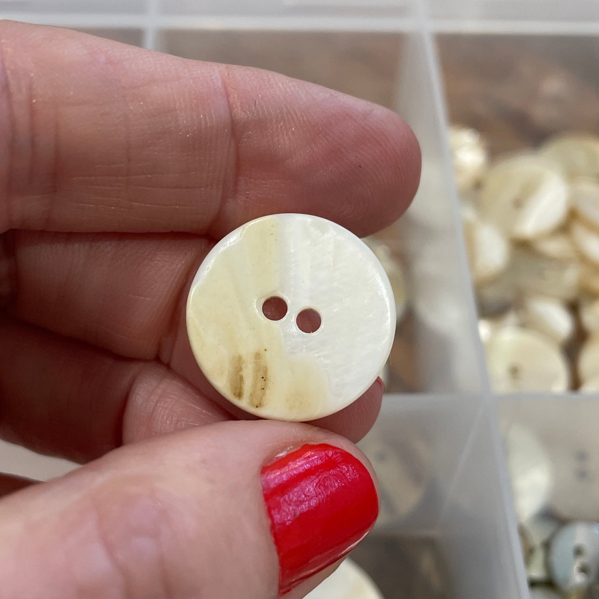 Hand holding a round button with two holes above a container of buttons.