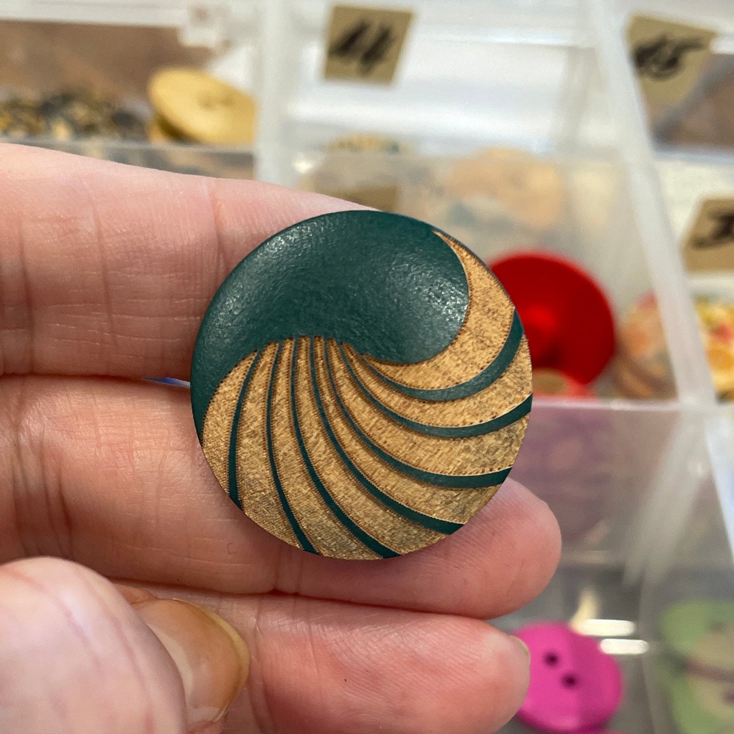 Hand holding a black and gold spiral-patterned button with a display of various buttons in the background.