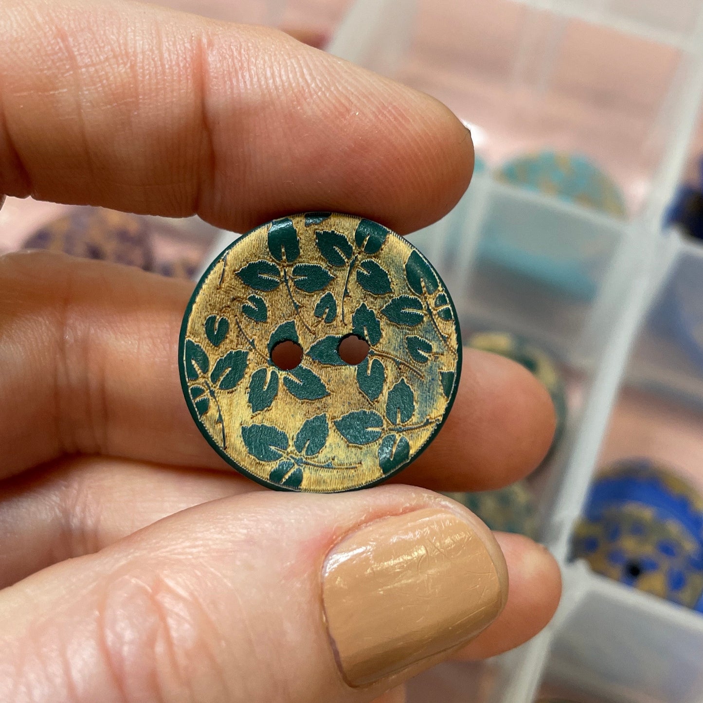 Hand holding a decorative button with intricate design, with a container of buttons in the background.
