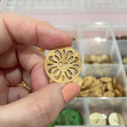 Hand holding a gold decorative object with a detailed pattern, with a container of similar objects in the background.