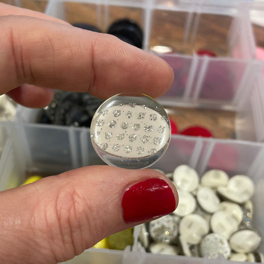 Hand holding a silver button with a clear container of buttons in the background
