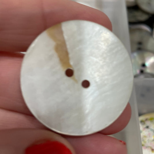 Close-up of a hand holding a white button with a central hole, with a blurred background of various buttons.