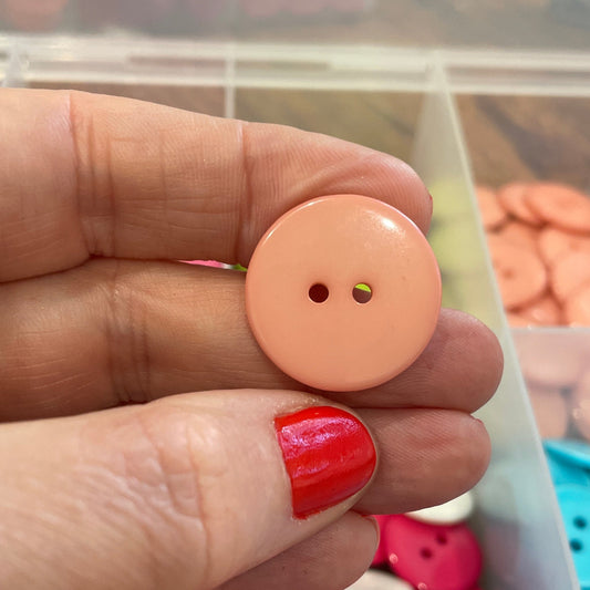 Hand holding a peach-colored button with a container of colorful buttons in the background