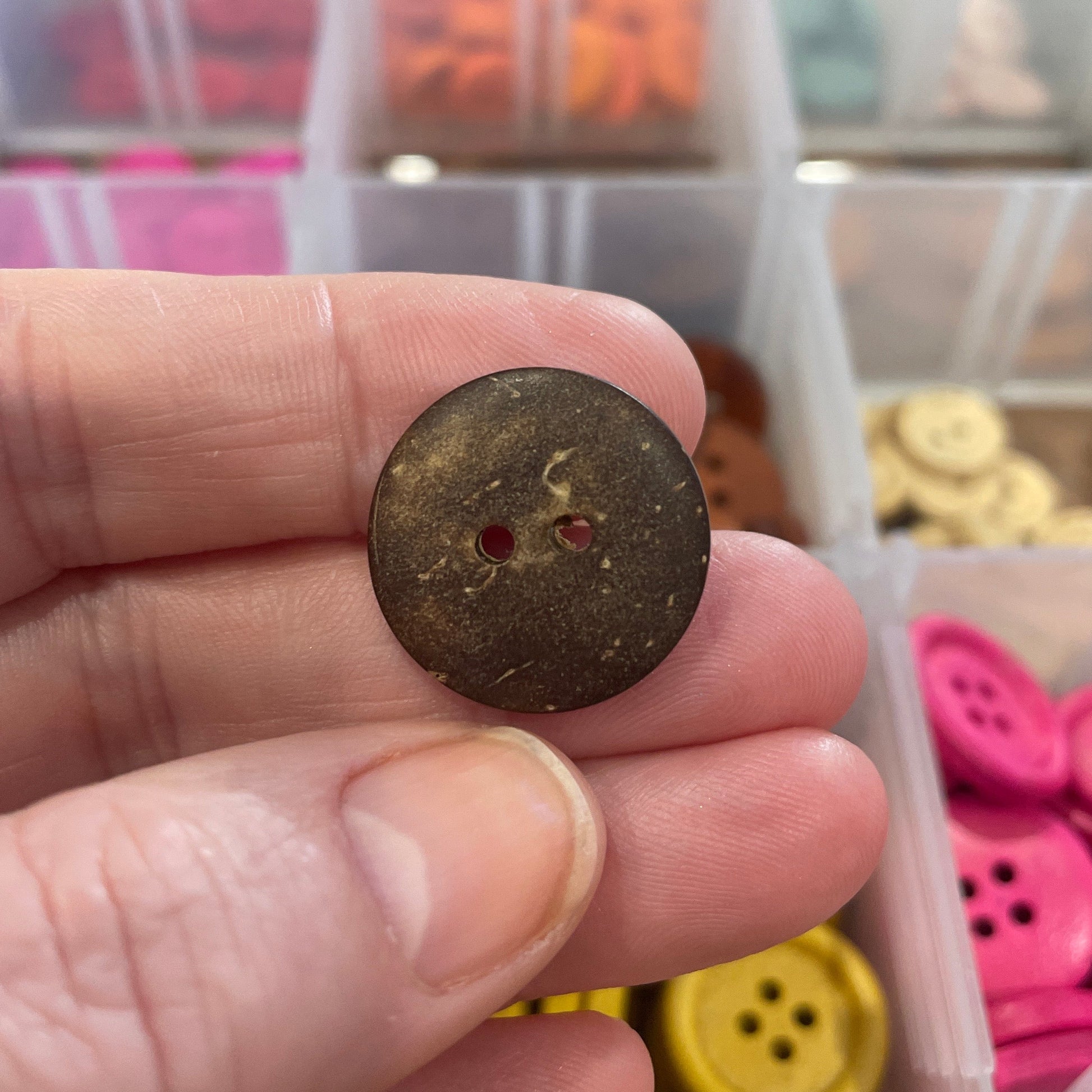 Hand holding a brown button with a container of colorful buttons in the background