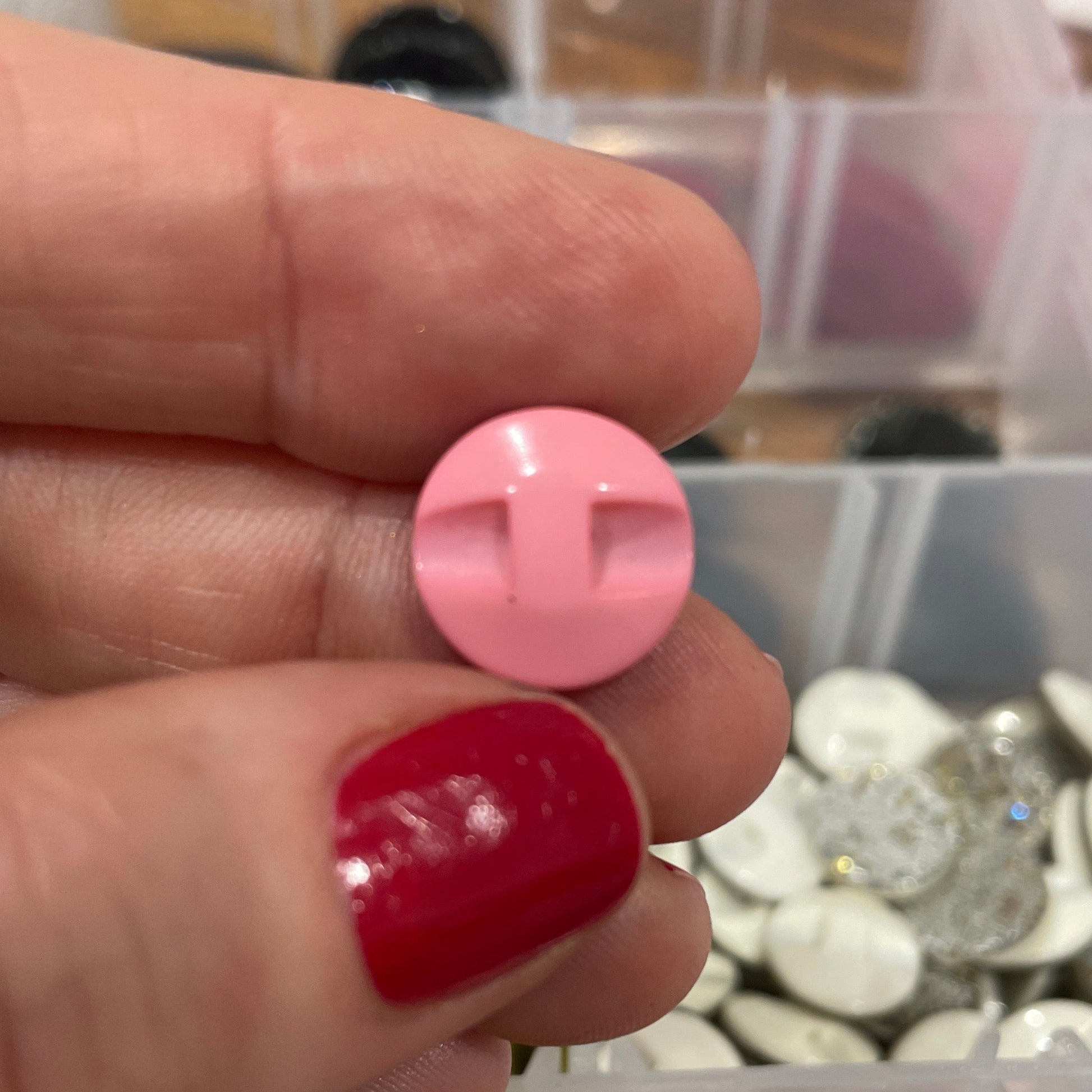 Hand holding a pink button with a blurred container of buttons in the background