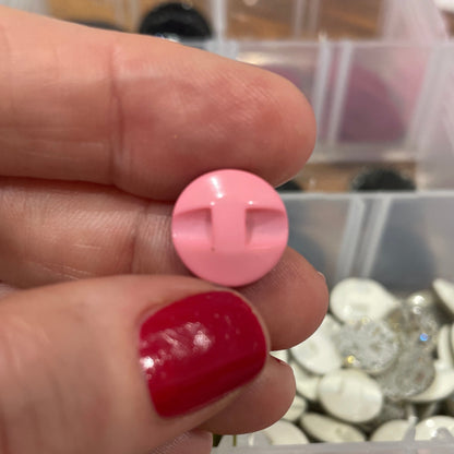 Hand holding a pink button with a blurred container of buttons in the background