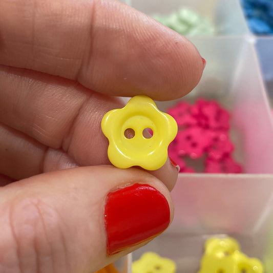 Hand holding a yellow flower-shaped button with a blurred background of a container with buttons.