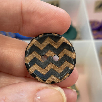 Hand holding a black and gold zigzag patterned button with a blurred background of a button collection.