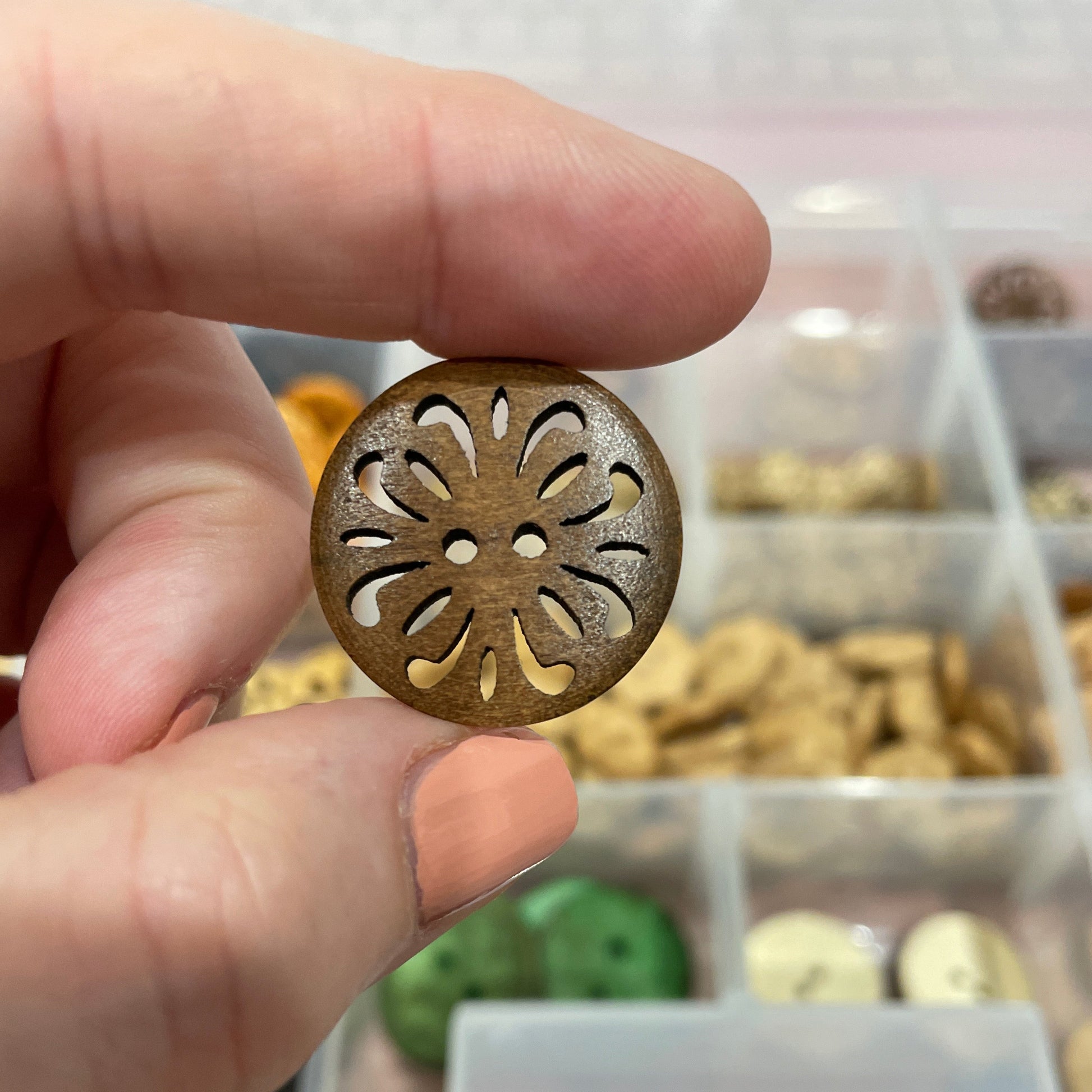Hand holding a decorative button with intricate design, blurred background of a container with buttons.
