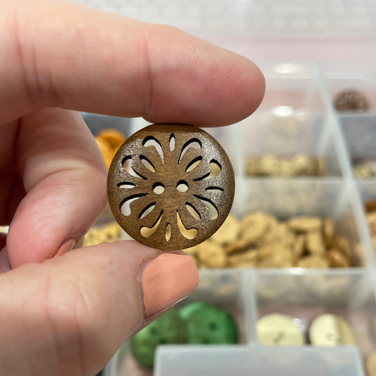 Hand holding a decorative button with intricate design, blurred background of a container with buttons.