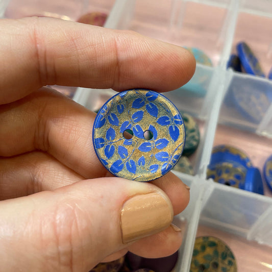 Hand holding a decorative button with a blue floral pattern, surrounded by a container of similar buttons.