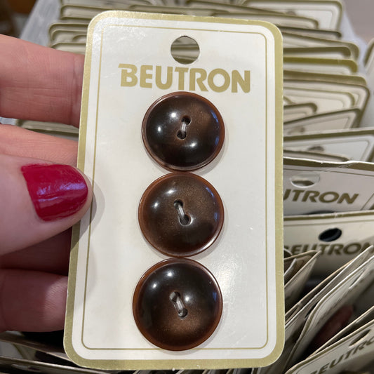 Three brown buttons on a Beutron card held by a hand with red nail polish.