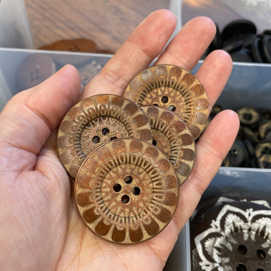 Hand holding three decorative buttons with intricate patterns, with a display of more buttons in the background.
